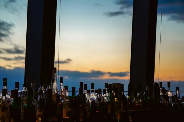 Silhouetted bottles along a bar in front of tall windows and a sunset sky