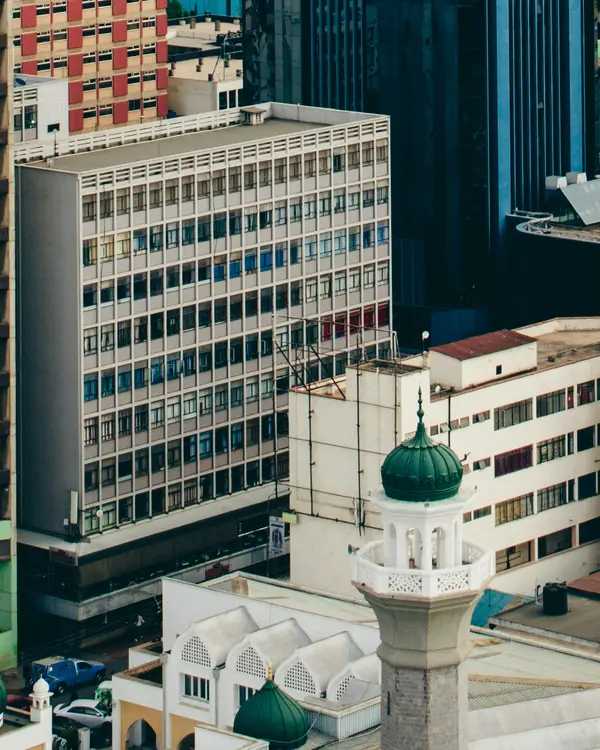 A white minaret with a green dome in front of a dark glass tower and older office blocks