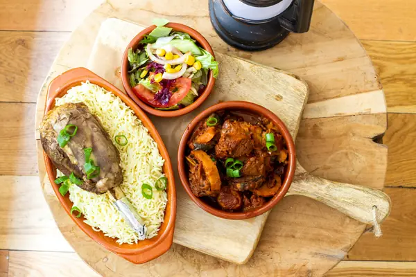 Clay dishes of rice with lamb, a salad and a stew on a wooden board