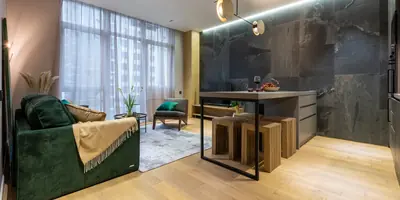 A studio flat with a green sofa, tall curtained windows and a small kitchen island against a dark stone wall (example photo, not of this place)