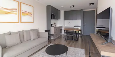 A bright living room with a grey sofa, a round dining table and a compact kitchen behind it (example photo, not of this place)