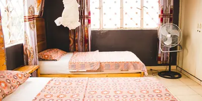 A sunny guest room with two single beds in patterned orange covers, printed curtains, a mosquito net and a standing fan (example photo, not of this place)
