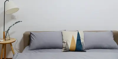A neatly made bed with grey linen and a patterned cushion beside a wooden side table and a floor lamp (example photo, not of this place)