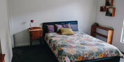 A double bed with a flowered quilt and purple pillows in a plain white room with wooden furniture (example photo, not of this place)