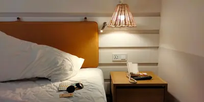 A bed with white linen against a mustard headboard, a woven lamp above a wooden bedside table with a telephone (example photo, not of this place)
