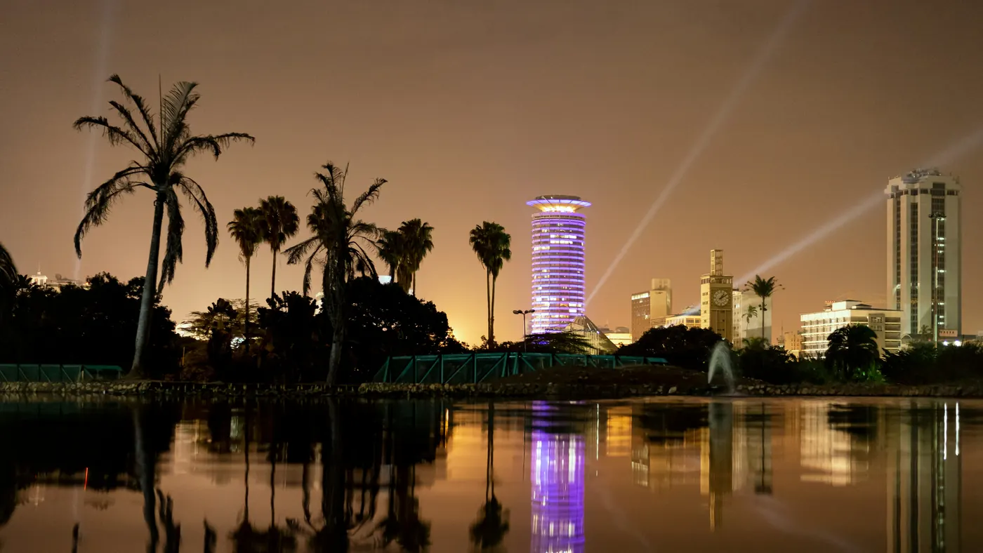 The KICC tower lit purple at night behind palm trees, reflected in a still lake in central Nairobi