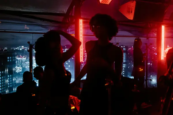 Silhouettes of people in a bar lit by red neon, city lights behind the windows