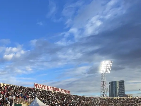 The Nyayo National Stadium name sign above a packed stand in Nairobi at golden hour, a floodlight shining against a blue sky