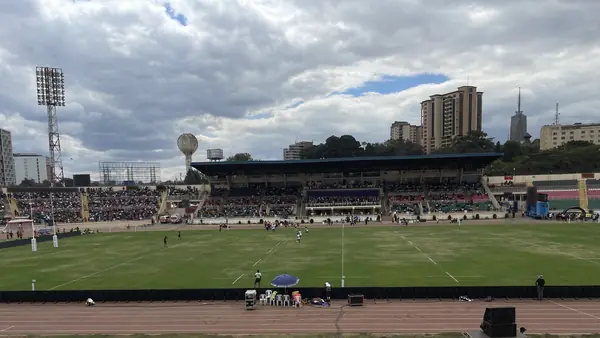 The main stand of Nyayo National Stadium with the Nairobi skyline and a round water tower behind it, players on the pitch