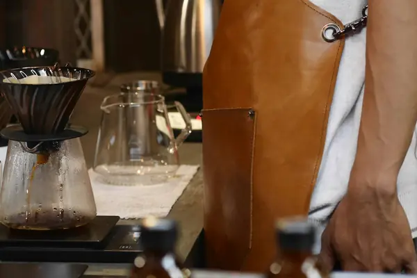 Pour-over coffee dripping into a glass jug on a scale beside a barista in a leather apron