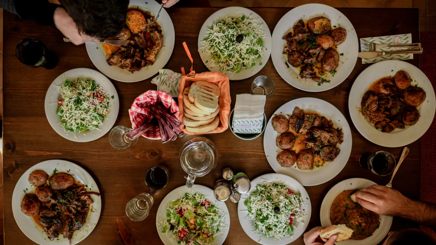 Overhead view of a wooden table set with plates of roast meat, potatoes, salads and bread shared by friends