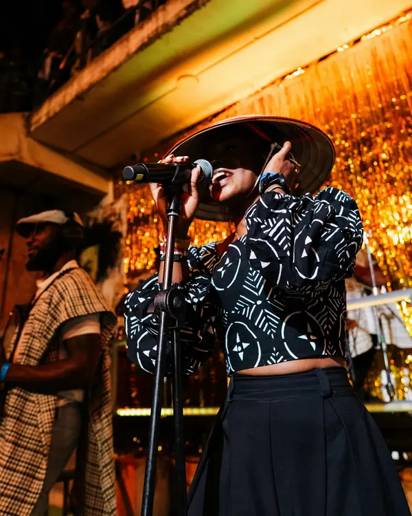 A singer in a wide hat and patterned top performing at a microphone against a gold curtain