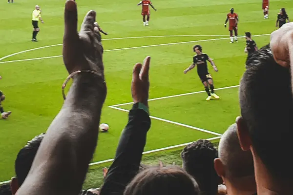 Fans' raised arms in the foreground as players in red and black move across a football pitch