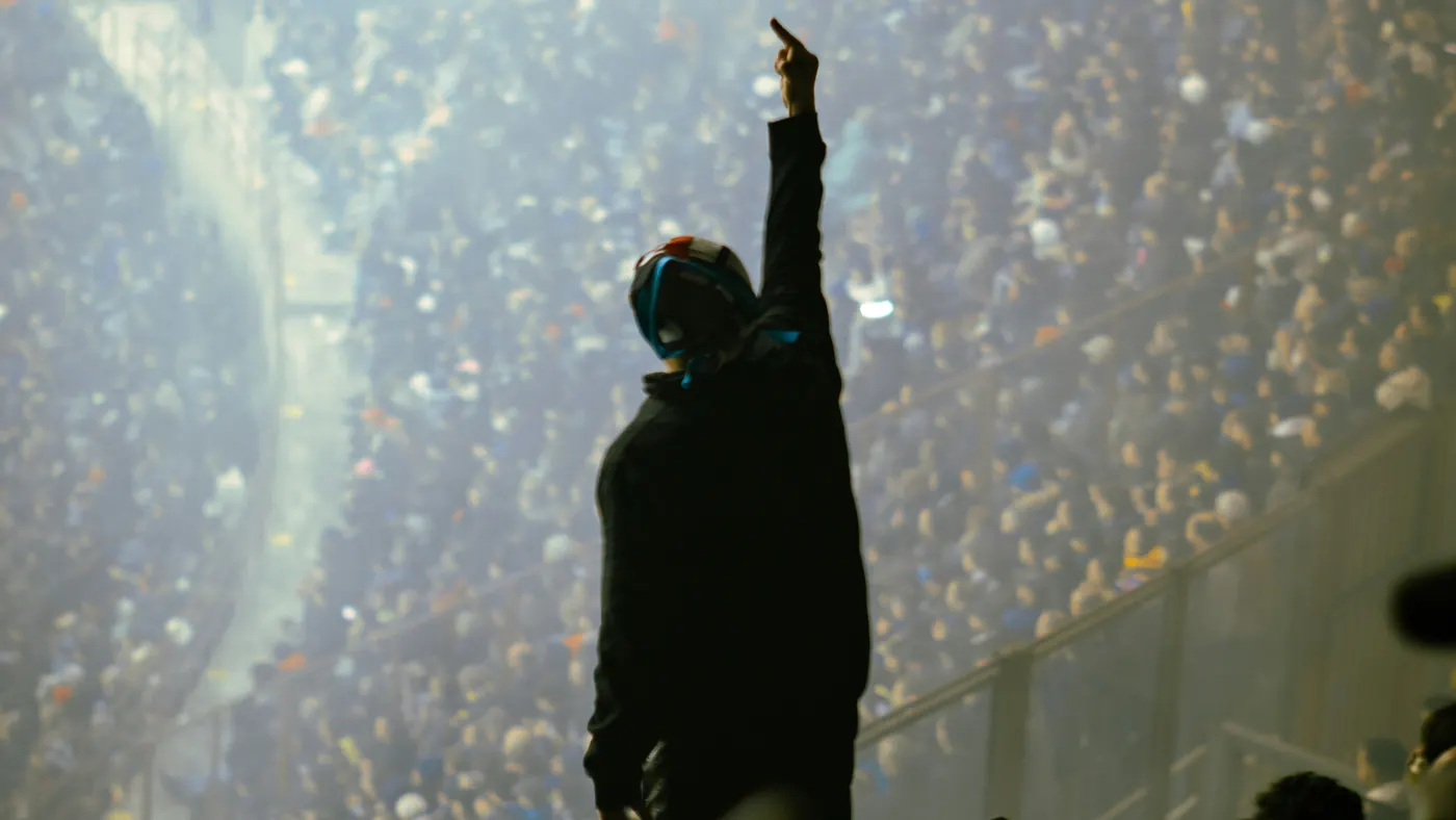 A fan silhouetted against a packed, smoky stadium stand, arm raised high