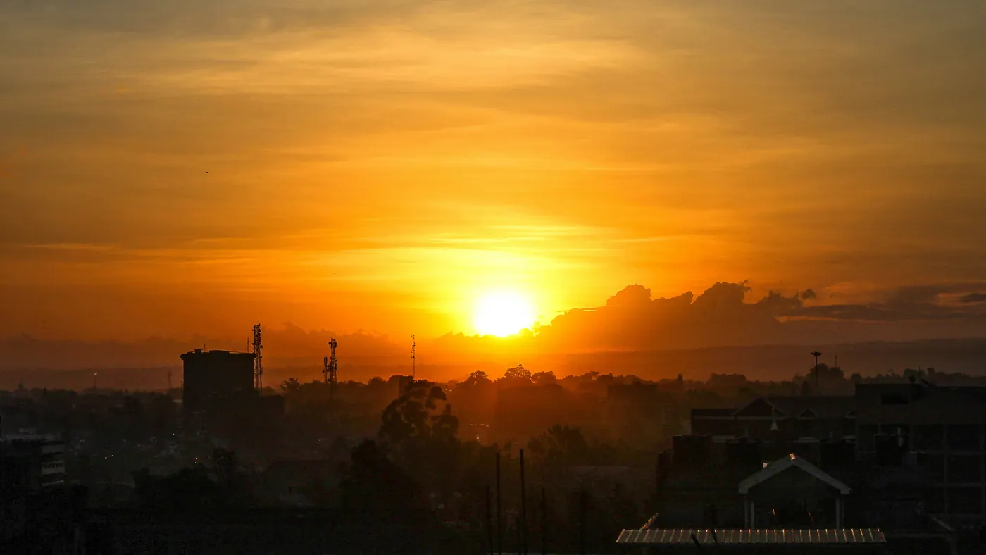 An orange sun setting over the silhouetted rooftops and trees of a city suburb